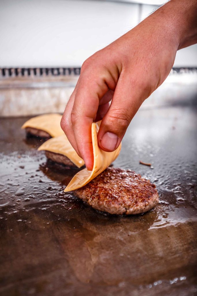 Main plaçant une tranche de fromage sur un steak cuit sur une plancha professionnelle.